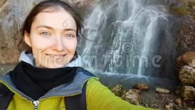 有趣的十几岁女孩做了一个有趣的自拍，而旅行的背景是一个美丽的瀑布。 她做鬼脸，做鬼脸。